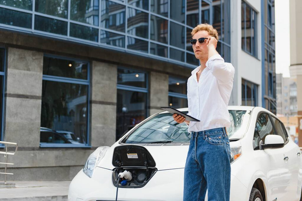 Business man standing near charging electric car and using tablet in the street. - Foto, Imagen