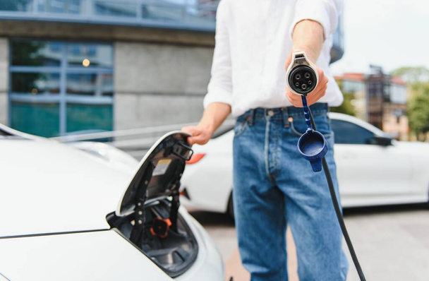Stylish man inserts plug of the charger into the socket of electric car close-up - Photo, image