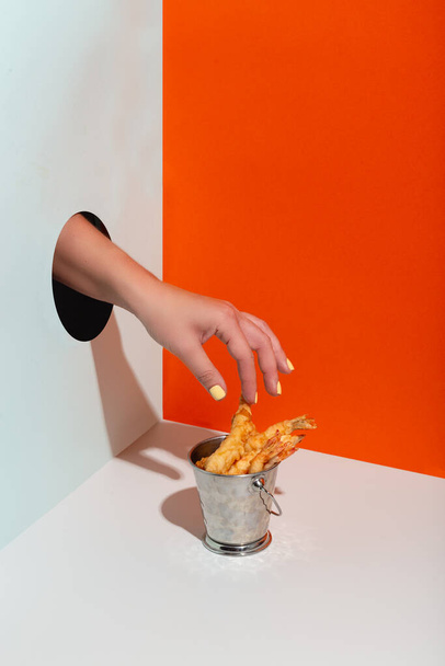 woman hand takes fried shrimp in metal bucket through paper hole modern hard shadow copy space - Fotó, kép