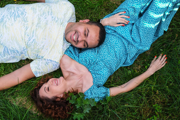 Loving couple lie on the grass look at each other and laugh. Man in a T-shirt and a curly woman in a blue dress on a romantic date. A Middle Eastern man with his caucasian wife. - Fotó, kép