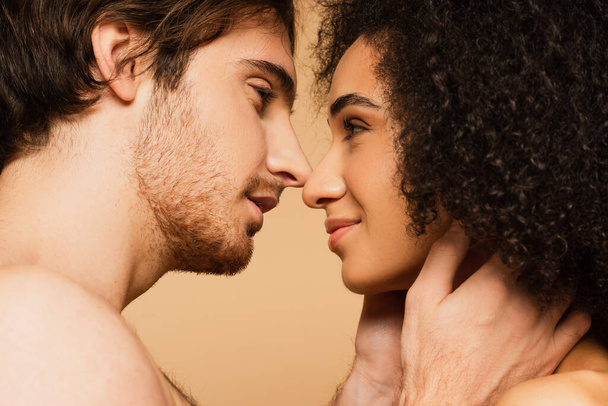 vista de cerca del hombre joven abrazando el cuello de la mujer hispana mientras está de pie cara a cara aislado en beige - Foto, Imagen