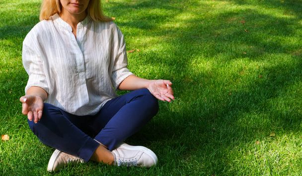 A woman in office casual clothes relaxes while sitting cross-legged on green grass in the shade of trees at lunchtime. Relax outdoors. A woman is meditating under the shade - Photo, Image
