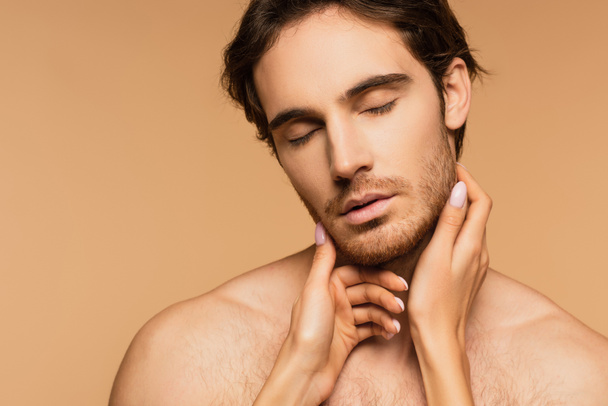 young shirtless man with closed eyes near female hands touching his face isolated on beige - Fotoğraf, Görsel