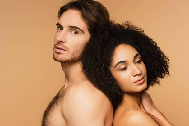 shirtless man looking at camera while standing back to back with sexy latin woman isolated on beige - 写真・画像