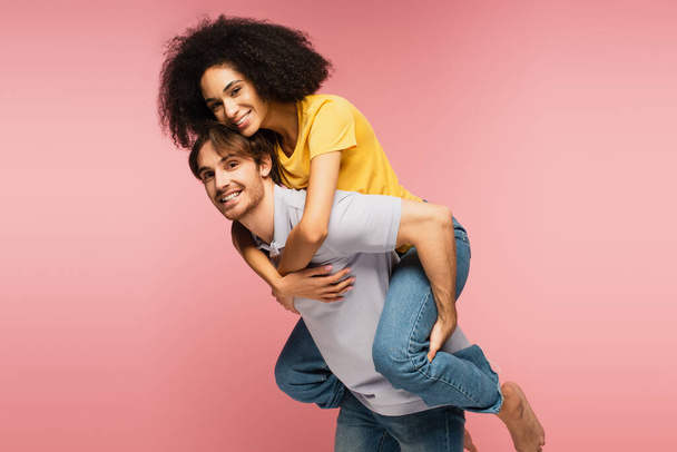 young and happy multiethnic couple smiling at camera while having fun isolated on pink - Photo, Image