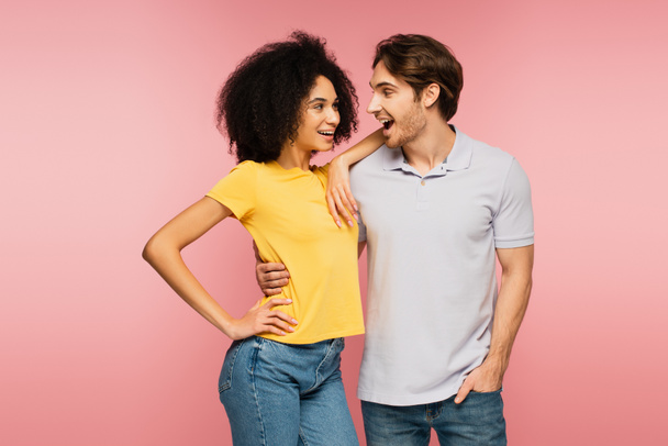 amazed man looking at joyful latin girlfriend standing with hand on hip isolated on pink - Photo, Image