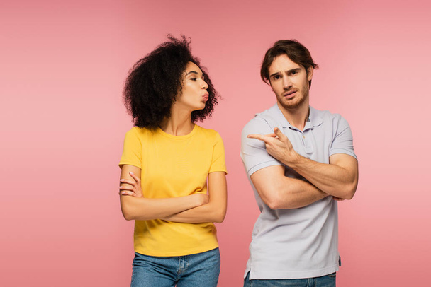 offended man pointing at latin girlfriend standing with crossed arms and grimacing isolated on pink - Foto, Imagen