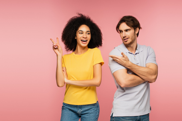 upset man pointing at excited hispanic woman showing victory gesture isolated on pink - Photo, Image