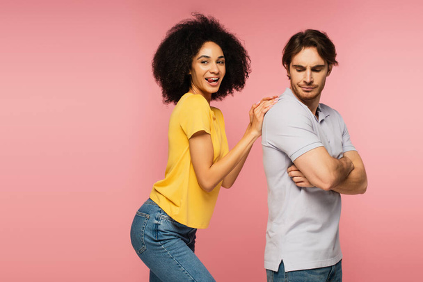 cheerful latin woman sticking out tongue while touching back of upset man standing with crossed arms isolated on pink - Фото, изображение