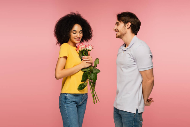 tyytyväinen latino nainen tilalla kimppu ruusuja lähellä nuori poikaystävä eristetty vaaleanpunainen - Valokuva, kuva