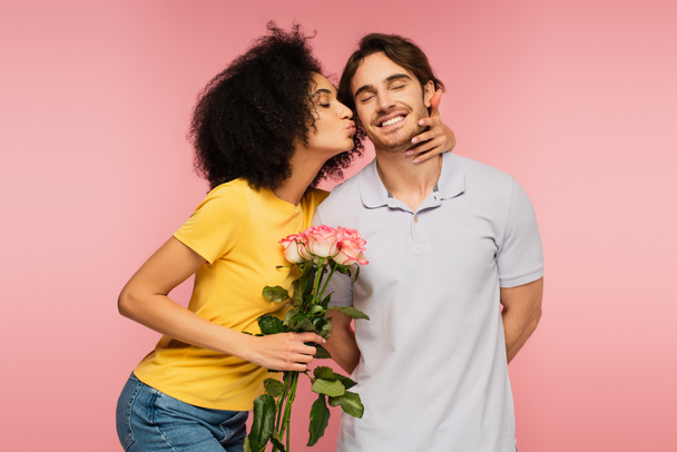 pretty latin woman with bouquet of roses kissing joyful man standing with closed eyes isolated on pink - Fotografie, Obrázek