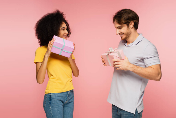 excited multiethnic couple smiling while holding gift boxes isolated on pink - Foto, imagen