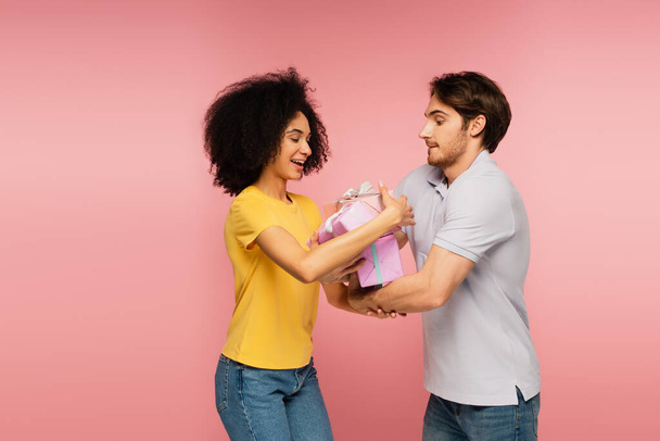 Молодой человек дарит множество подарков изумленной латинской подружке, изолированной на розовом - Фото, изображение