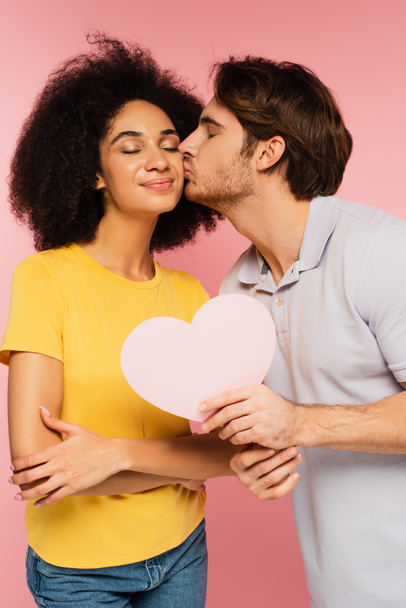 man with paper cut heart kissing happy hispanic girlfriend isolated on pink - Фото, зображення
