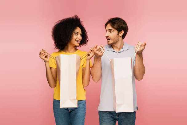 alegre pareja interracial mirándose entre sí mientras se abren bolsas de compras aisladas en rosa - Foto, Imagen