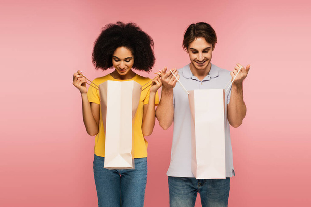 Zufriedenes multiethnisches Paar blickt isoliert auf rosa Einkaufstüten - Foto, Bild