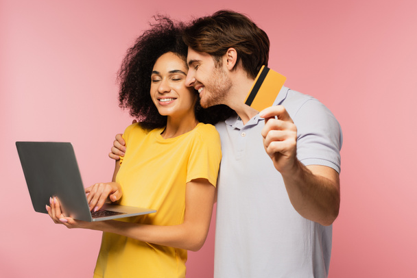 hombre alegre con tarjeta de crédito abrazando novia hispana feliz usando el ordenador portátil aislado en rosa - Foto, imagen