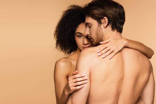seductive latin woman looking at camera while hugging sexy shirtless man isolated on beige - Foto, Imagem