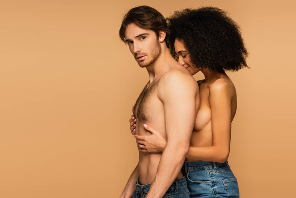 topless latin woman with closed eyes hugging muscular torso of shirtless man looking at camera isolated on beige - Photo, Image