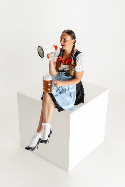 Young beautiful woman, waitress in traditional Austrian or Bavarian costume sitting with megaphone on huge box isolated over white background. - Fotó, kép