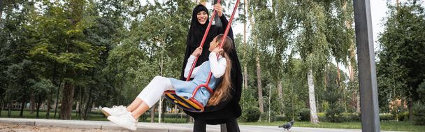 Аравійська жінка грає з дочкою на гойдалці на відкритому повітрі, на прапорі  - Фото, зображення