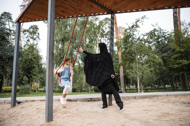 Muslim mother standing near child on swing in park  - Photo, Image