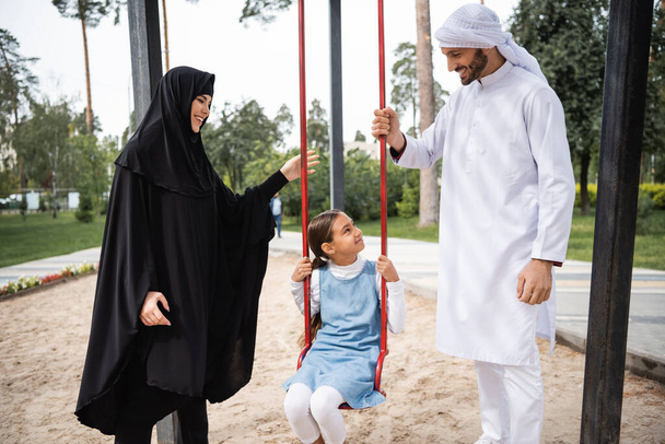 Smiling arabian parents looking at daughter on swing in park  - Valokuva, kuva
