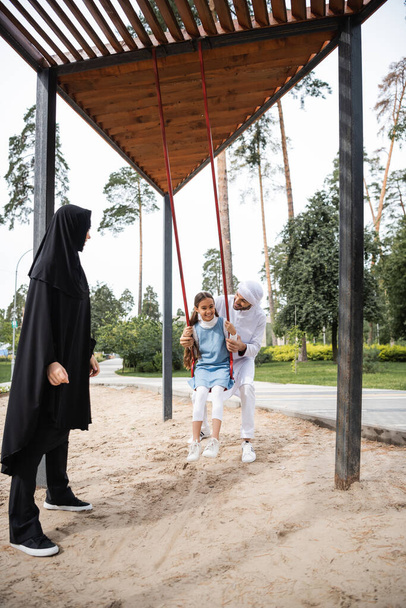 Μουσουλμάνος πατέρας περνάει χρόνο με την κόρη του σε κούνια και γυναίκα στο πάρκο  - Φωτογραφία, εικόνα