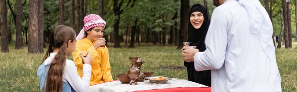 Улыбающаяся арабка, сидящая рядом с мужем, дети и чай в парке, баннер  - Фото, изображение
