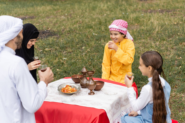 Happy muslim kids holding tea in glasses near parents in park  - Foto, afbeelding