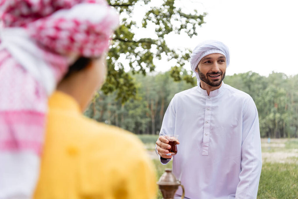 Smiling muslim man holding glass of tea near blurred son outdoors  - 写真・画像
