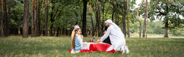 公園で家族とピクニック中にお茶を保持するイスラム教徒の父,バナー  - 写真・画像