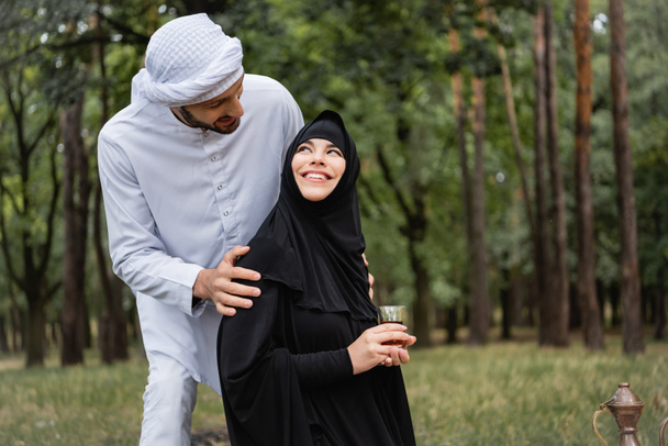Арабский мужчина в традиционной одежде обнимает жену чаем в парке  - Фото, изображение