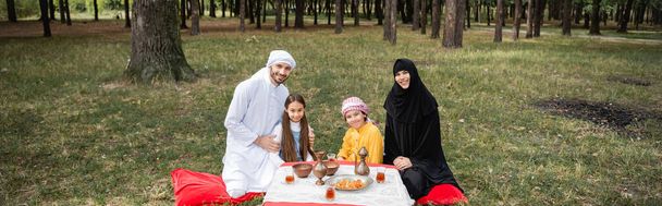 Glimlachende moslim familie kijkt naar camera in de buurt van voedsel in het park, banner  - Foto, afbeelding