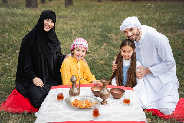 Sorridenti genitori arabi che abbracciano i bambini vicino al cibo e al tè nel parco  - Foto, immagini