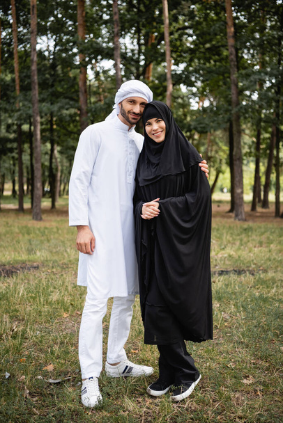 Μουσουλμάνος άνδρας σε kufiya αγκαλιάζει χαμογελαστή γυναίκα στο πάρκο  - Φωτογραφία, εικόνα