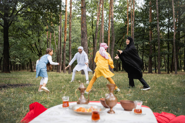 Família muçulmana feliz em roupas tradicionais jogando perto de chá desfocado no parque  - Foto, Imagem