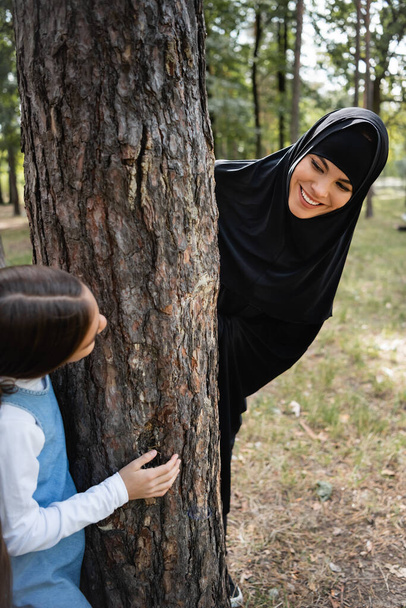 Muslim mother in hijab looking at daughter near tree in park  - 写真・画像