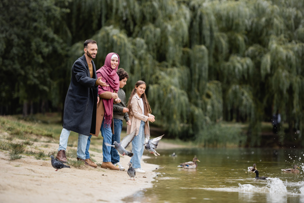 Cheerful muslim man pointing at birds on lake near family in park  - 写真・画像