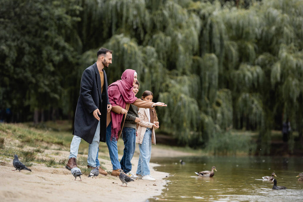 Smiling muslim mother pointing at birds on lake near husband and kids on beach - Photo, Image