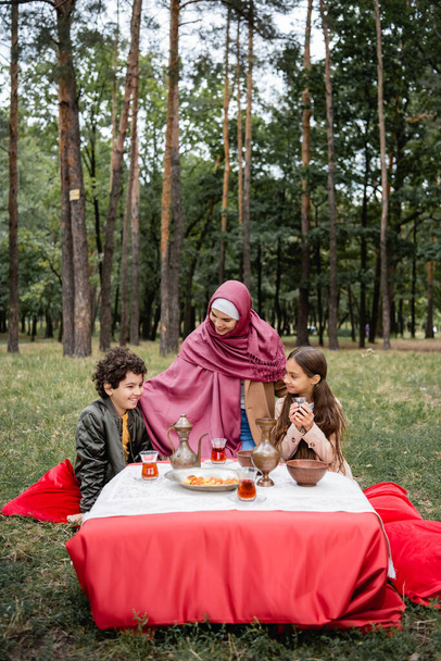 Madre árabe positiva abrazando a los niños cerca del té y las frutas secas en el parque  - Foto, imagen