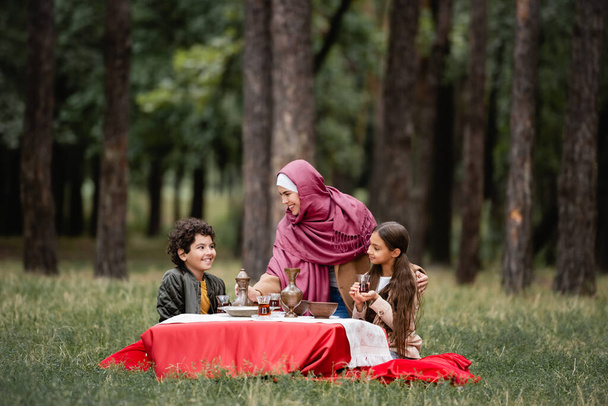 Arabian woman smiling at kids with tea in traditional glasses in park  - Photo, Image