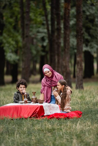Smiling kids holing tea in glasses near muslim mother in hijab in park  - Photo, Image