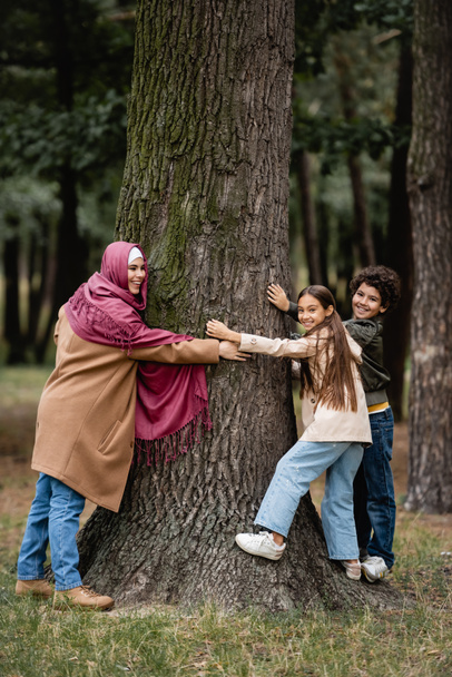 Cheerful muslim woman and children hugging tree in park - Фото, изображение