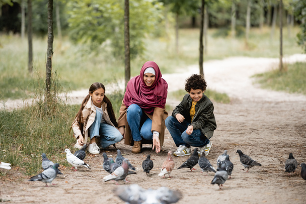 Positive arabian mother and children feeding blurred doves in park  - Foto, immagini