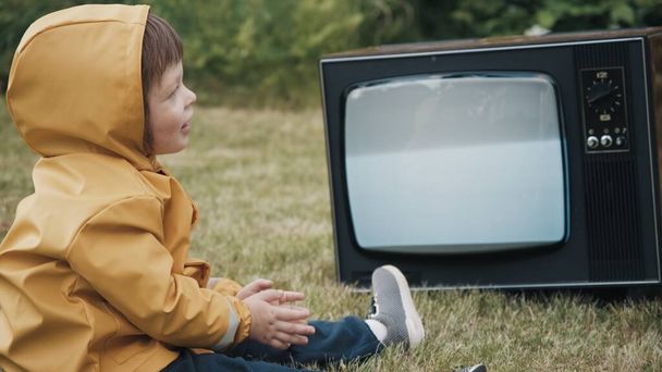Child dressed in raincoat is watching retro TV. Baby claps his hands, rejoices - Foto, Imagen