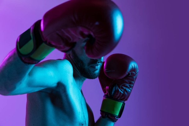 Un entraînement professionnel de boxeur masculin isolé sur fond violet dans la lumière au néon. Attaque adverse. Poinçonnage - Photo, image