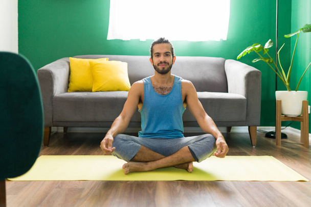 Handsome young man preparing to meditate in his living room. Portrait of a happy man doing yoga while looking at the camera - Foto, imagen