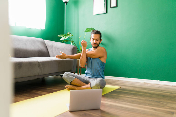 Βλέποντας ένα βίντεο φροντιστήριο. Ευτυχισμένος νεαρός άνδρας κάνει ασκήσεις τεντώματος, ενώ μετά από ένα online σπίτι βίντεο προπόνηση - Φωτογραφία, εικόνα