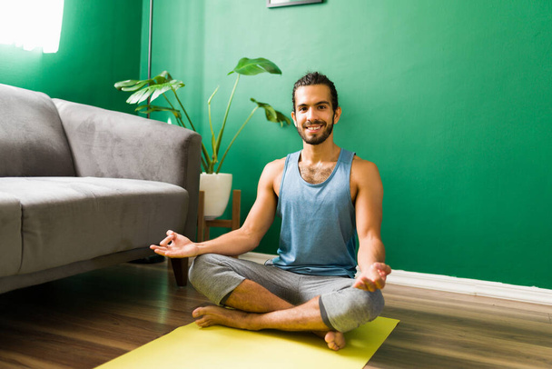 Porträt eines entspannten hispanischen Mannes in einer lockeren Yoga-Pose, der Blickkontakt aufnimmt und lächelt, bevor er seine Meditation beginnt  - Foto, Bild